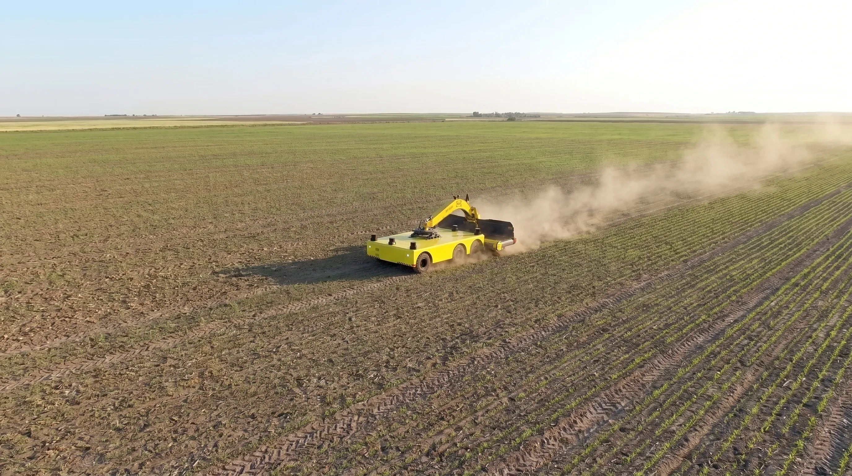 Aerial view of a yellow electric platform transporter working across an open field with an implement mounted on its deck, raising dust behind it, with drilled rows and wheel tracks in the soil.